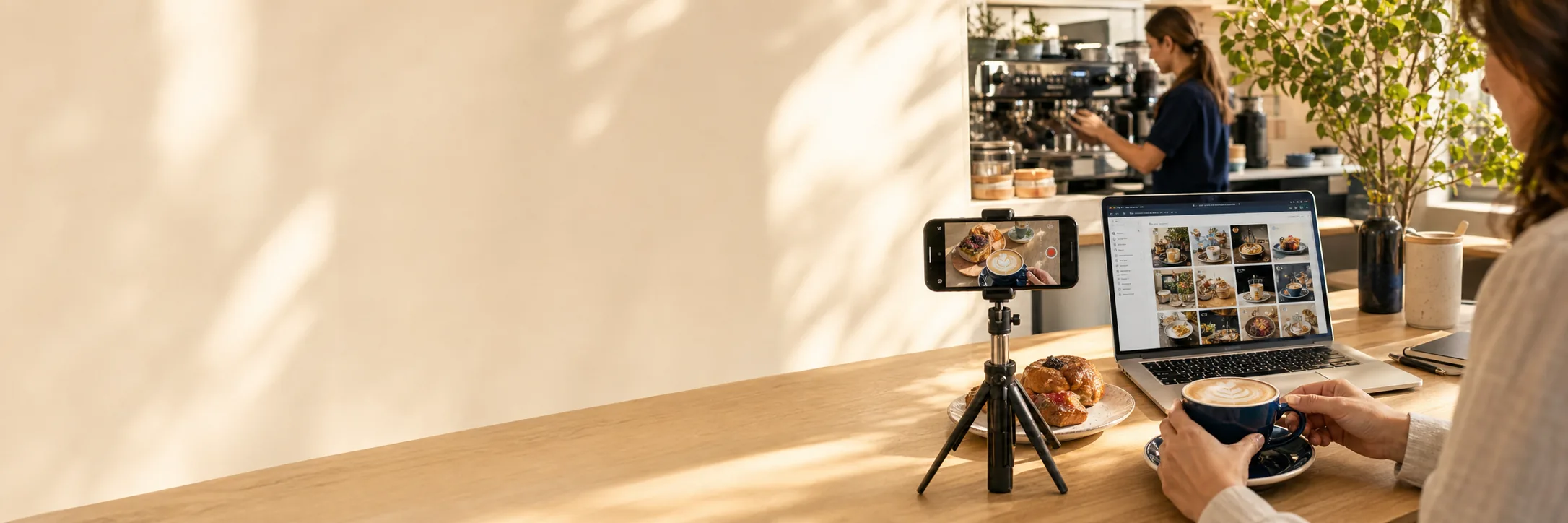 A phone on a small tripod filming a latte at a café counter, with a laptop showing a grid of social posts